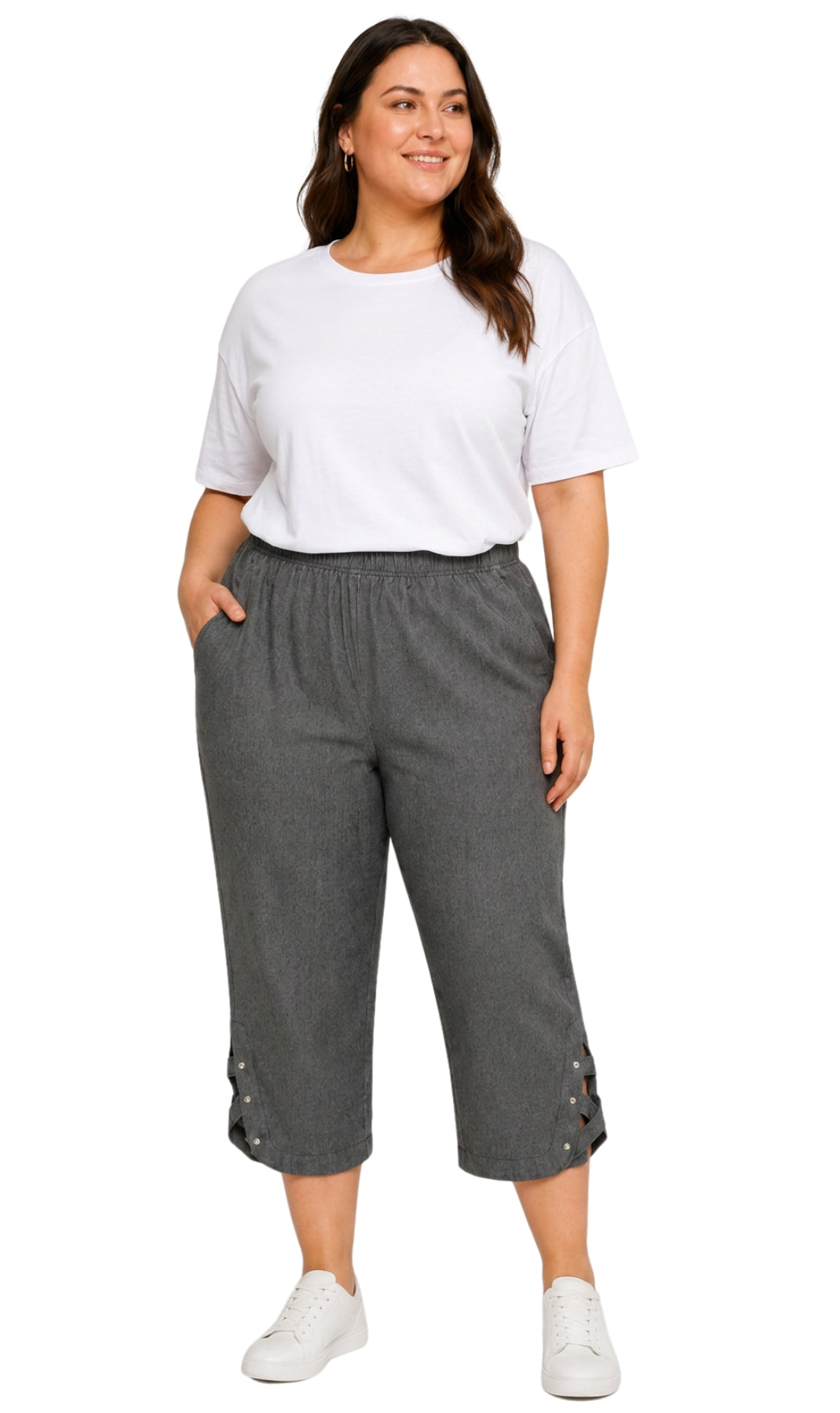 A woman modeling a pair of dark gray cropped pants with decorative side buttons, paired with a white t-shirt and white sneakers, against a plain white background.